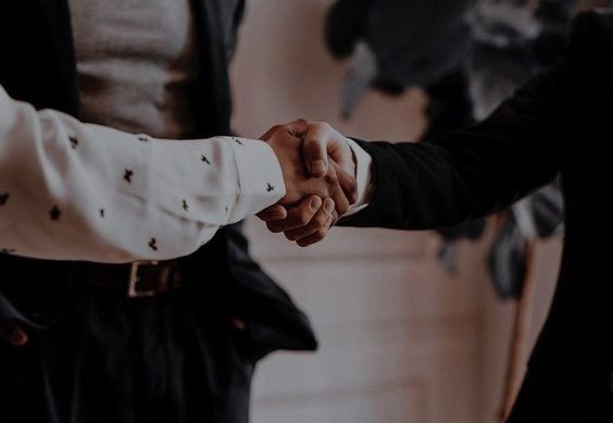 Partnership Handshake Two people shaking hands in a professional setting. The person on the left is wearing a white shirt with a pattern, and the person on the right is wearing a black suit.