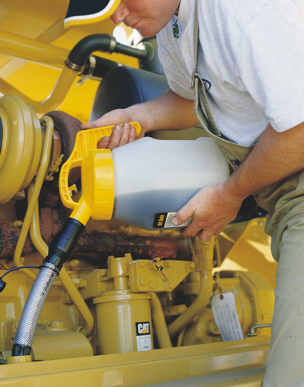 story-hero-1 A person wearing a cap is refueling a yellow construction vehicle. They are kneeling and using a hose to transfer fluid from an Oilsafe drum container into the vehicle's engine compartment.